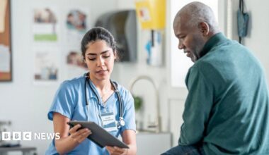 A nurse in blue scrubs with a stethoscope consults with a Black patient in a clinic, using a tablet. Warm, professional healthcare scene highlighting patient care and medical teamwork.