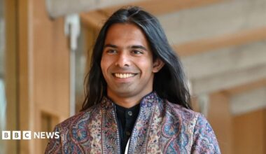 A person wearing a patterned jacket, with long flowing dark hair smiles as they walk towards the camera in the Scottish Parliament building