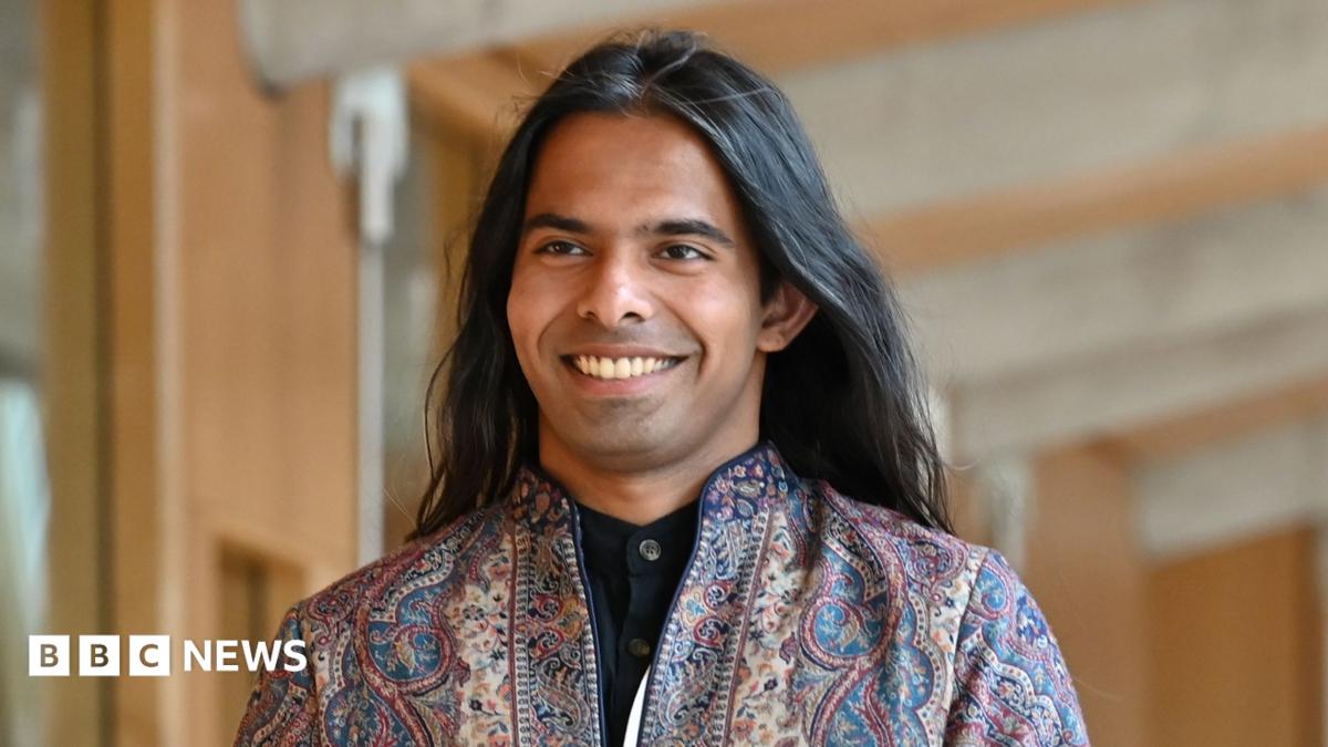 A person wearing a patterned jacket, with long flowing dark hair smiles as they walk towards the camera in the Scottish Parliament building