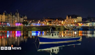 A blue fishing boat sits on calm water in Oban Harbour at night, with colourful lights from waterfront buildings reflecting across the water beneath a dark sky.