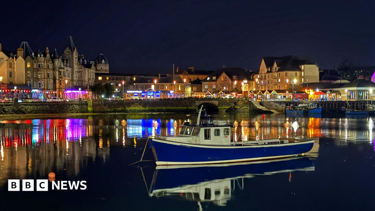 A blue fishing boat sits on calm water in Oban Harbour at night, with colourful lights from waterfront buildings reflecting across the water beneath a dark sky.