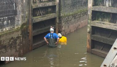 Two people stand in shallow water inside a canal lock wearing white safety helmets. One is in a blue T-shirt and chest waders, the other in a yellow high-visibility jacket. The lock's wooden gates and blackened brick walls rise steeply on either side.