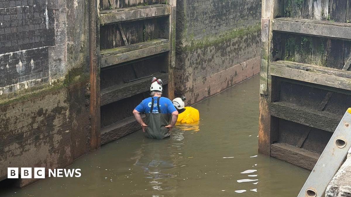 Two people stand in shallow water inside a canal lock wearing white safety helmets. One is in a blue T-shirt and chest waders, the other in a yellow high-visibility jacket. The lock's wooden gates and blackened brick walls rise steeply on either side.
