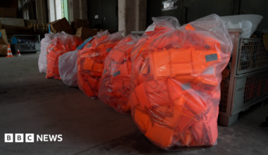 Large numbers of bright orange lifejackets are seen in plastic bags inside a warehouse