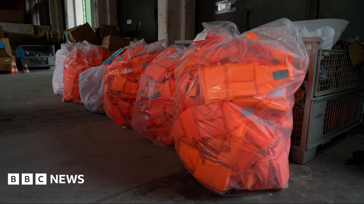 Large numbers of bright orange lifejackets are seen in plastic bags inside a warehouse