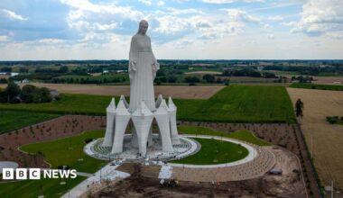 A statue of the Virgin Mary rises out of green and yellow fields in Poland on a cloudy sky.