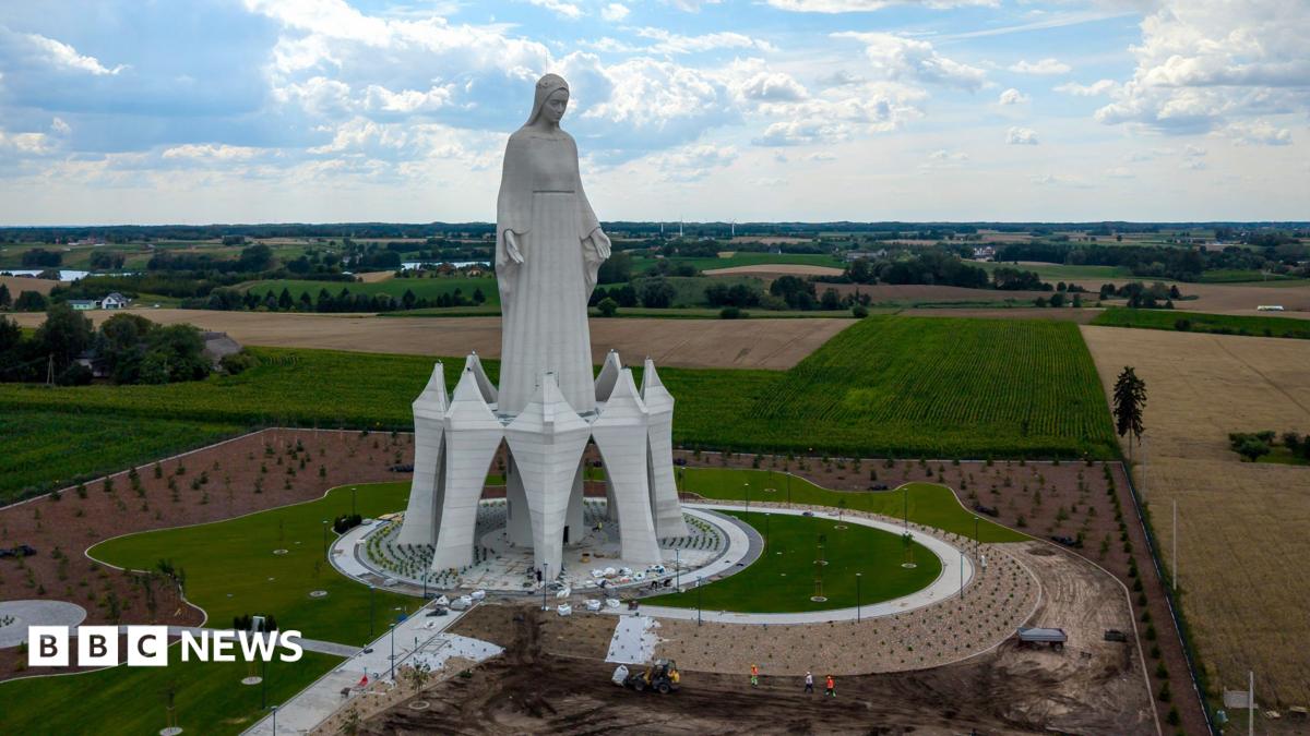 A statue of the Virgin Mary rises out of green and yellow fields in Poland on a cloudy sky.