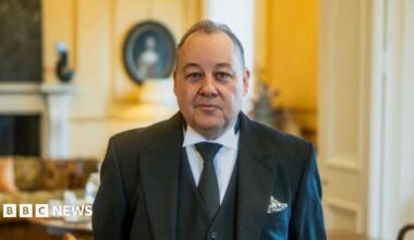 A butler smiles at the camera as he stands in a large drawing room. He has short dark hair and wears a black suit jacket, black waistcoat, white shirt and green tie.