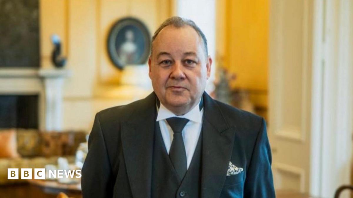 A butler smiles at the camera as he stands in a large drawing room. He has short dark hair and wears a black suit jacket, black waistcoat, white shirt and green tie.