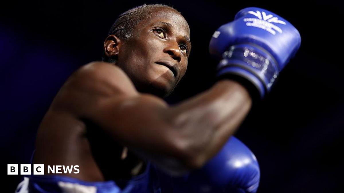 Nuhu Batte, with blue boxing gloves on and with an arm flexed, preparing to fight.