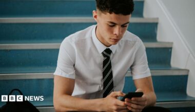 an adolescent boy in a short-sleeved shirt and a tie sitting on stairs, looking at a phone