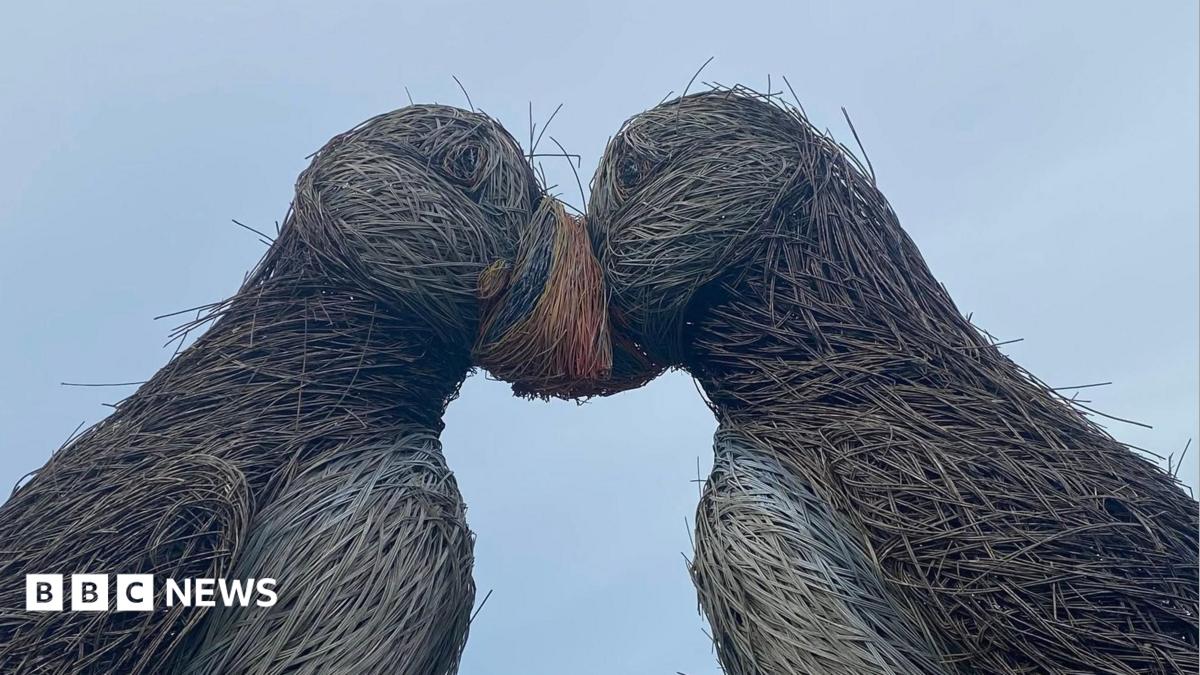 A side on shot of the two puffin sculptures with their heads together. The willow is frayed and coming off in many places and the paintwork is faded. A grey-ish sky is visible in the background.