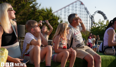 Image shows a family sitting on an artificial grass verge watching the solar eclipse, and wearing eclipse glasses. The outline of the Big One rollercoaster can be seen in the background.