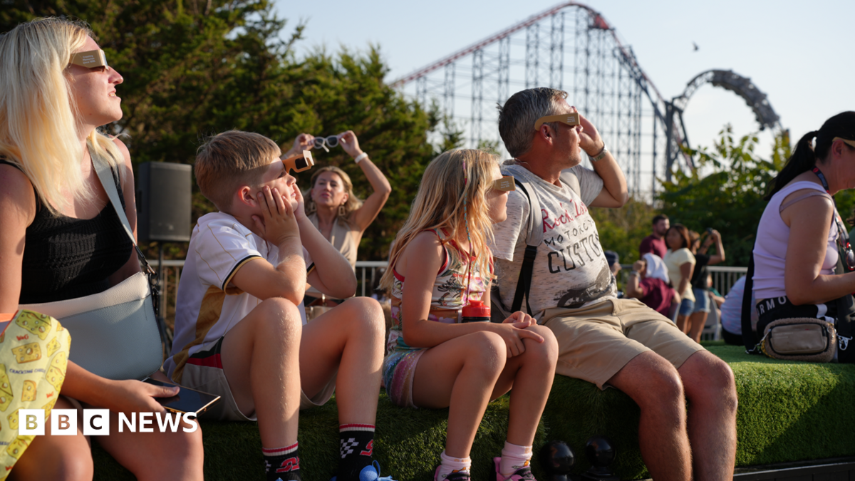 Image shows a family sitting on an artificial grass verge watching the solar eclipse, and wearing eclipse glasses. The outline of the Big One rollercoaster can be seen in the background.