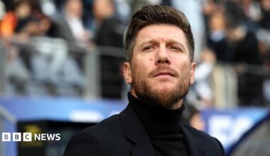 A man wearing a black polo neck and blazer with a quiffed hairstyle and a beard. He is standing in front of a stand full of football fans and is gazing into the distance.
