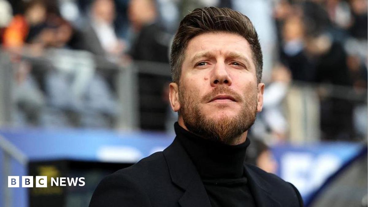 A man wearing a black polo neck and blazer with a quiffed hairstyle and a beard. He is standing in front of a stand full of football fans and is gazing into the distance.