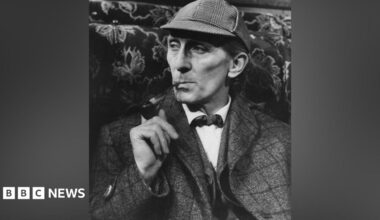 A black and white photo of a man in a cap and tweed jacket. He is smoking a pipe.