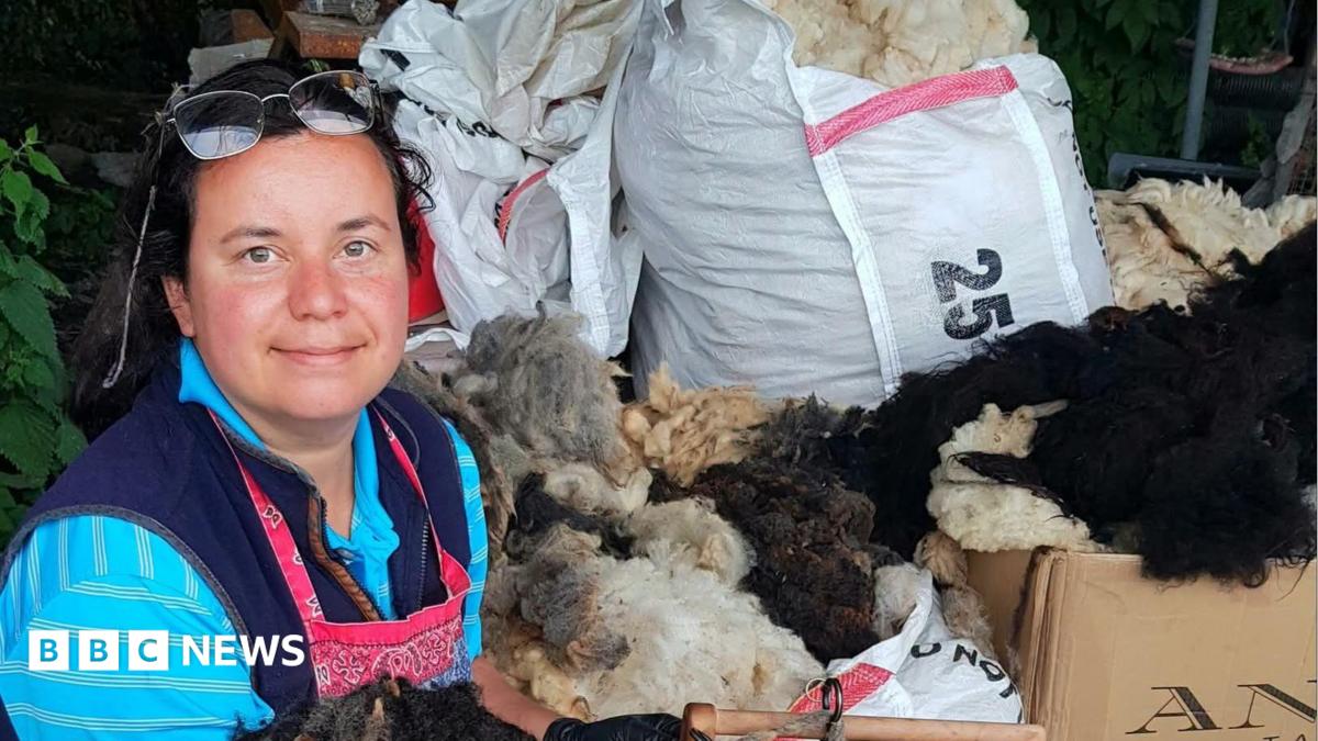 A woman in her late 30s with dark hair tied back. She has glasses on her head and is wearing a blue striped shirt with a pink apron on top. She is surrounded by bags of wool.