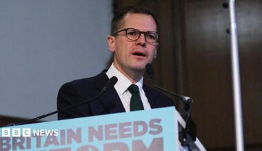 Headshot of Jenrick who speaks into two small microphones on stands on a podium in front of him that has a blue sign partially cut out of the frame, but you can read it says 'Britain needs reform'.