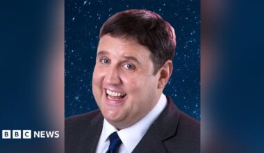 Close up of comedian Peter Kay with brown hair wearing a dark grey  suit and navy tie. He is standing in front of a background of navy blue and glitter. He is smiling.