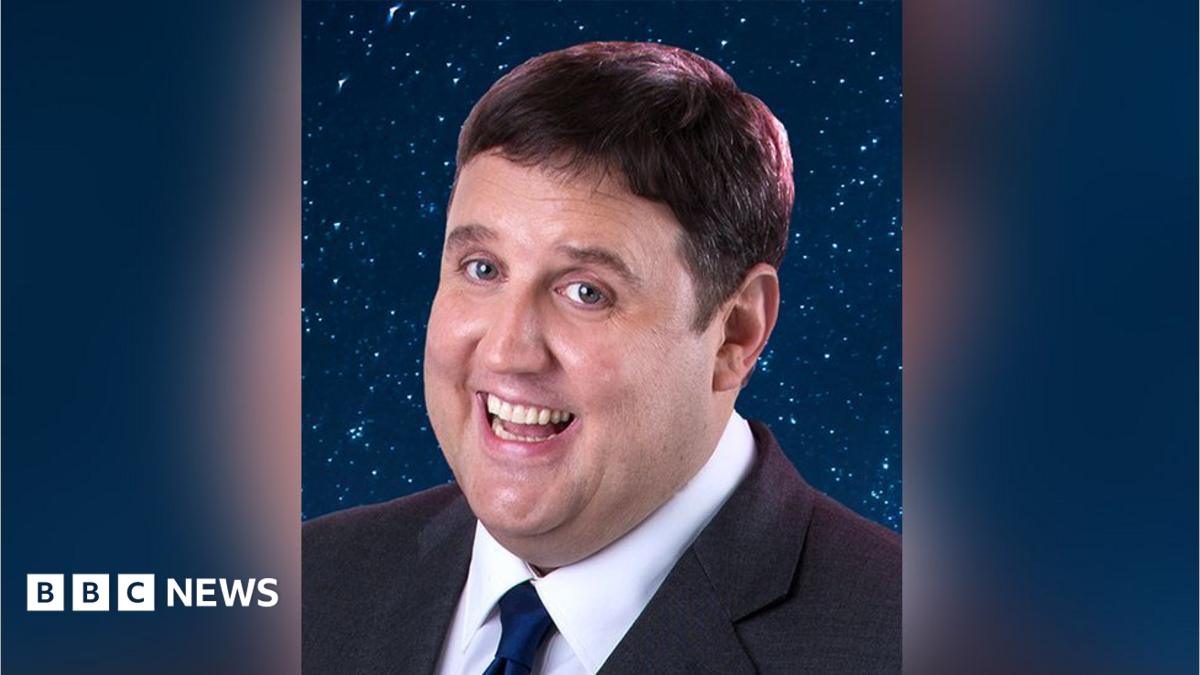 Close up of comedian Peter Kay with brown hair wearing a dark grey  suit and navy tie. He is standing in front of a background of navy blue and glitter. He is smiling.