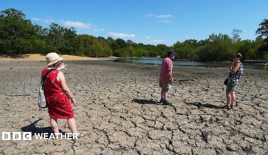 Three people standing on cracked earth of a dried out mud bed of Hollow Pond in East London on 11th August 2026