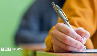 A close-up stock image of a young person's hand holding a silver pen while writing on paper on a desk. They are wearing a yellow sweatshirt. Behind, a blurred background shows another person sitting at a desk in a darker top, and behind them a green wall.