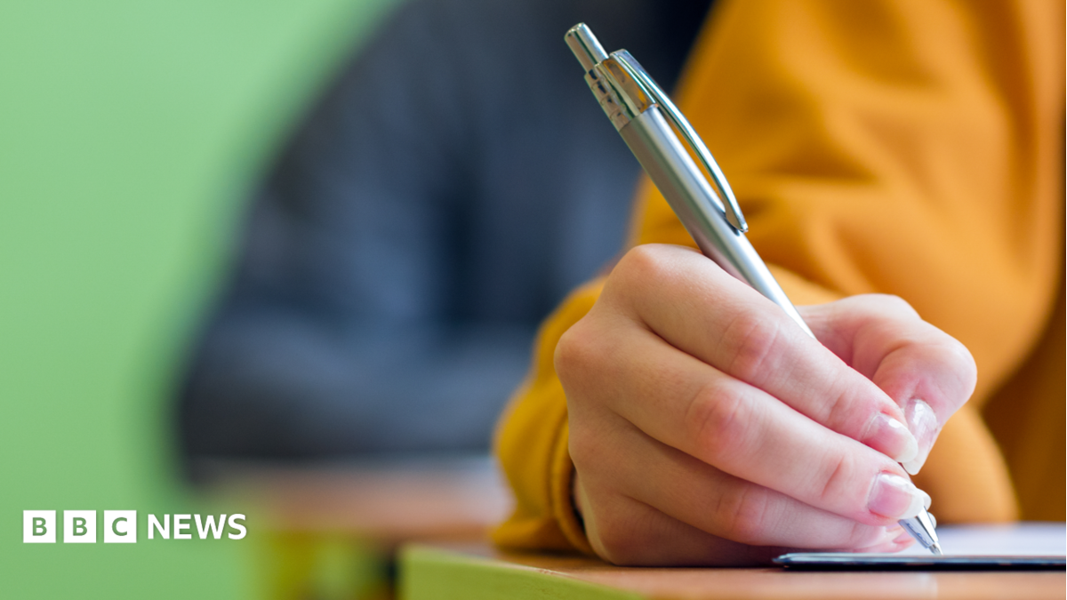 A close-up stock image of a young person's hand holding a silver pen while writing on paper on a desk. They are wearing a yellow sweatshirt. Behind, a blurred background shows another person sitting at a desk in a darker top, and behind them a green wall.