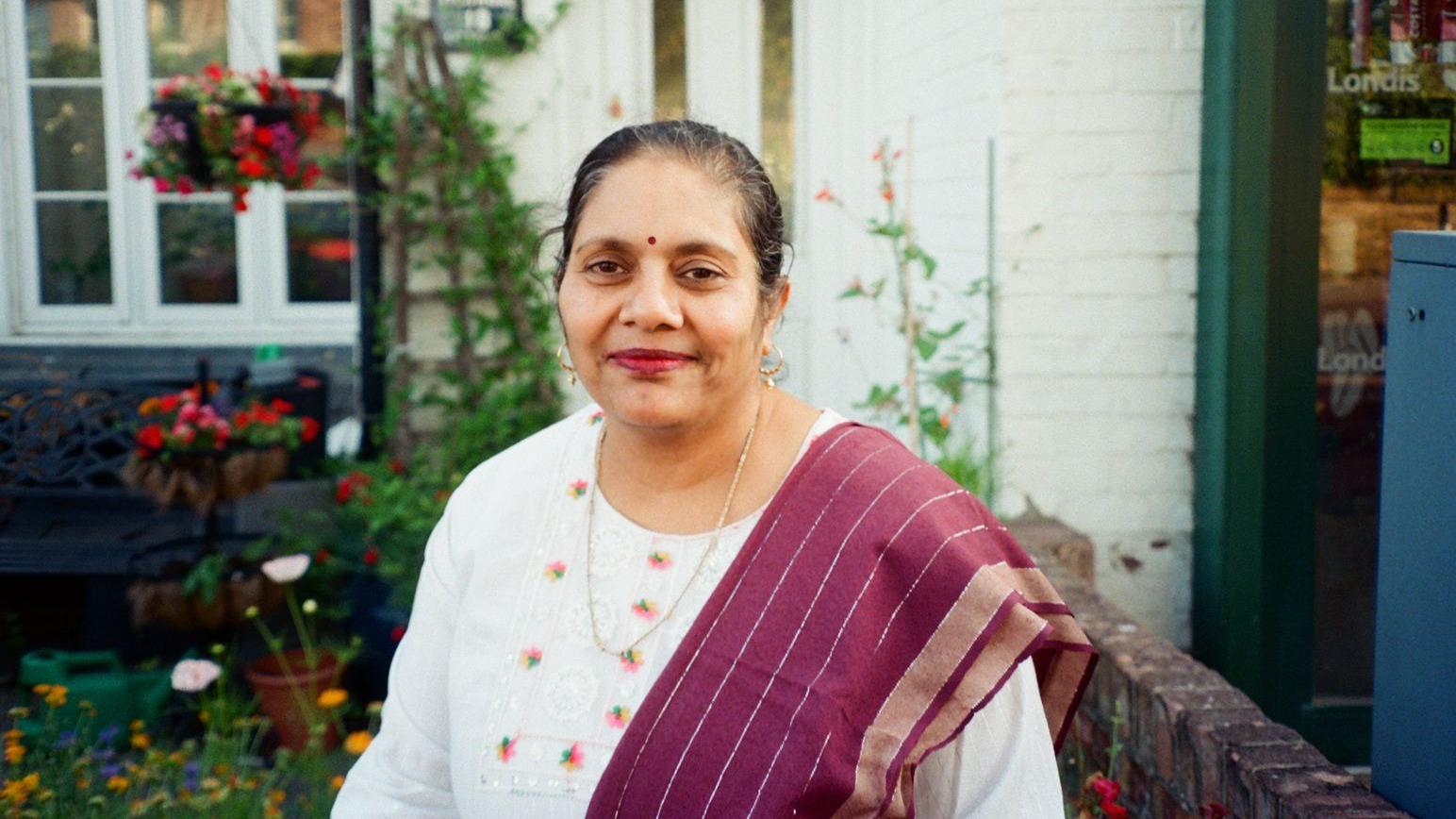 Jigna smiles in front of a garden next to her Londis shop. She has dark hair tied back and wears a white top with pink flowers embroidered on it, a long gold chain and a dark red scarf over one shoulder.