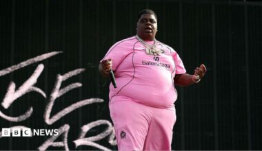 The rapper wearing a pink outfit with 'balenciaga' written on the chest, and several necklaces, holds a microphone in his extended right hand away against a black background with some chalk-like letters to his right