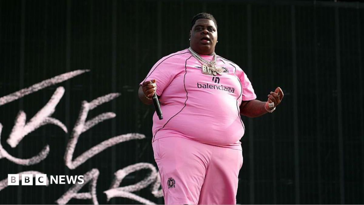 The rapper wearing a pink outfit with 'balenciaga' written on the chest, and several necklaces, holds a microphone in his extended right hand away against a black background with some chalk-like letters to his right
