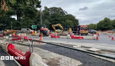 Roadworks at a large road junction, with excavators, construction workers in high-visibility clothing, cones and red-and-white barriers surrounding excavated sections of roadway. Newly laid tarmac and unfinished kerbs are visible in the foreground, while cars, a contractor van and a green motorway direction sign stand against a backdrop of trees under a cloudy sky.