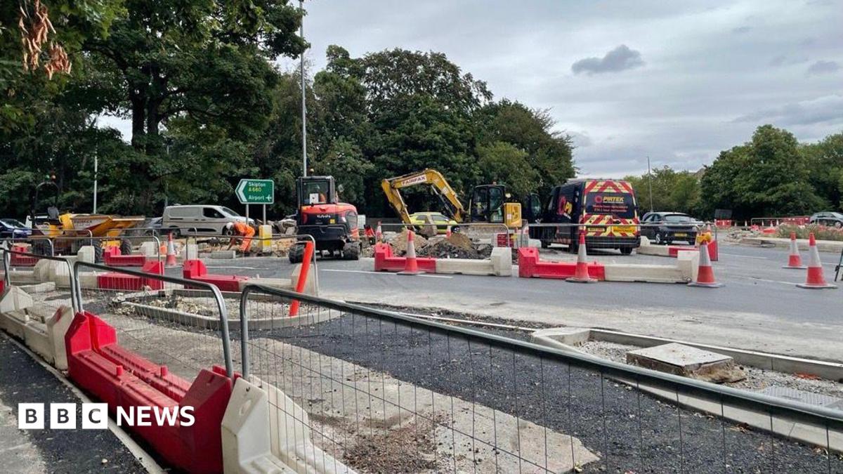 Roadworks at a large road junction, with excavators, construction workers in high-visibility clothing, cones and red-and-white barriers surrounding excavated sections of roadway. Newly laid tarmac and unfinished kerbs are visible in the foreground, while cars, a contractor van and a green motorway direction sign stand against a backdrop of trees under a cloudy sky.