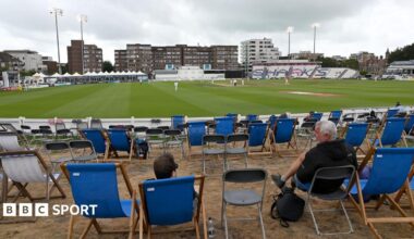 A view of the County Ground in Hove looking on to the pitch with two man sat in deckchairs, with several other empty deckchairs around them, under murky skies while the floodlights are on