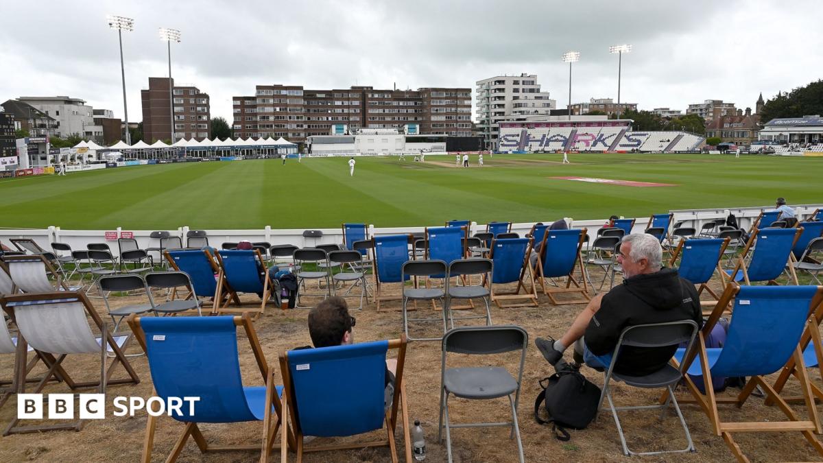 A view of the County Ground in Hove looking on to the pitch with two man sat in deckchairs, with several other empty deckchairs around them, under murky skies while the floodlights are on