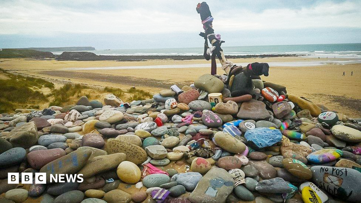 A pile of rocks and pebbles with messages scrawled in colourful writing on them on a beach. A wooden cross sits at the top of the pile.