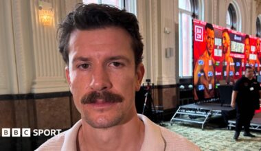 Boxer Nathan Heaney, who has dark hair and a moustache, stands to the side of a promotional dais advertising his next bout. A security man stands in the background to his left