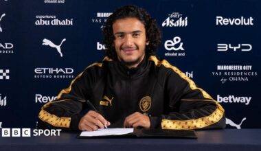 Ayyoub Bouaddi in a Manchester City tracksuit top holding a pen with a contract on the desk in front of him
