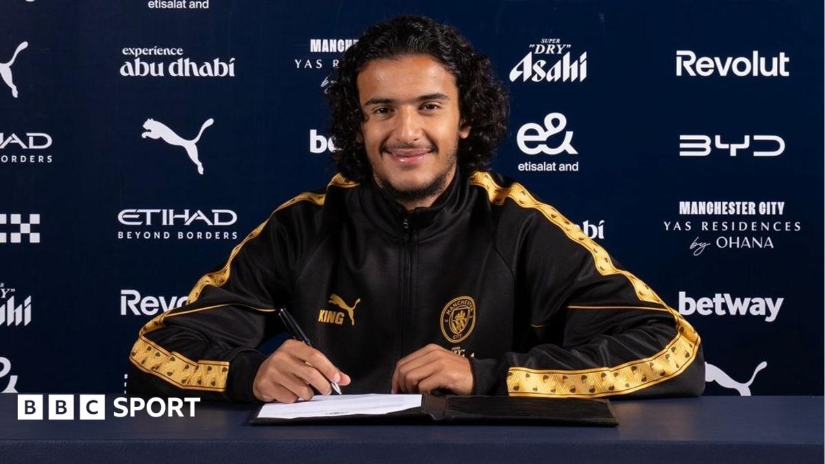 Ayyoub Bouaddi in a Manchester City tracksuit top holding a pen with a contract on the desk in front of him