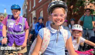 A girl with a bike helmet and school rucksack with others on bikes in the background.