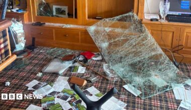 Significant damage to a wooden display cabinet following an apparent impact, with shattered glass panels, debris scattered across the floor, and contents displaced throughout the room.