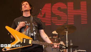 Tim Wheeler of Ash wears a dark Kurt Cobain T-shirt and a pale wood guitar. He is on stage with a drummer behind him and the word ASH in red on a black screen behind him