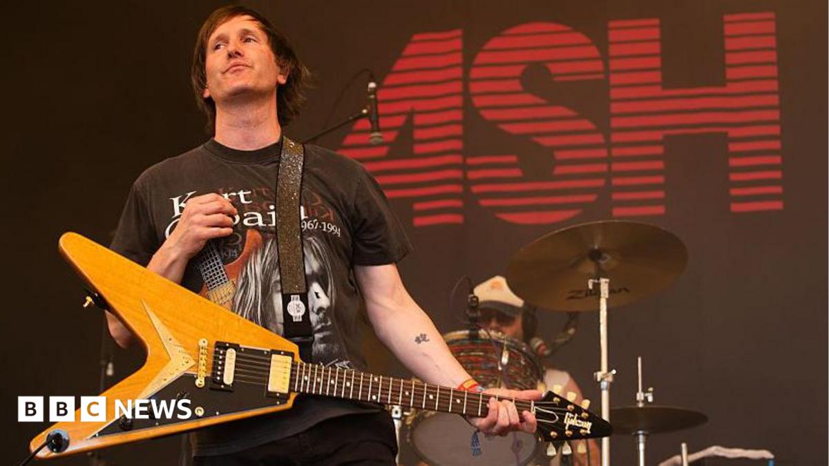 Tim Wheeler of Ash wears a dark Kurt Cobain T-shirt and a pale wood guitar. He is on stage with a drummer behind him and the word ASH in red on a black screen behind him