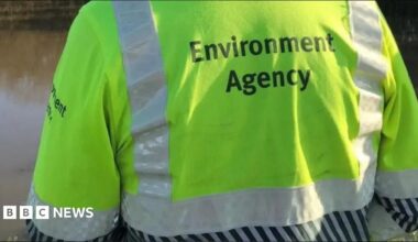 The back of an Environment Agency worker wearing a fluorescent yellow high-visibility jacket with reflective strips and the words “Environment Agency” printed on it.