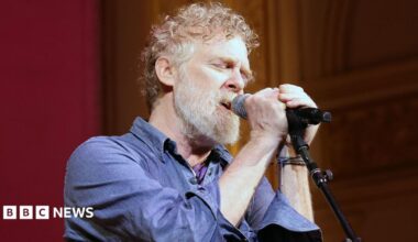 Glen Hansard has short curly reddish/brown/grey hair and a greyr beard and is singing with two hands on the microphone and his eyes closed