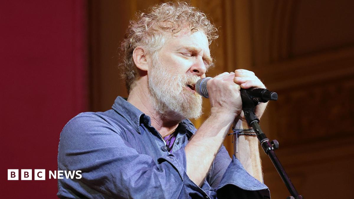 Glen Hansard has short curly reddish/brown/grey hair and a greyr beard and is singing with two hands on the microphone and his eyes closed