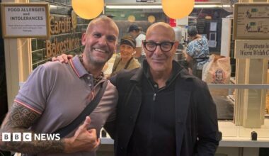 Owner of the Welsh cake store stood next to actor Stanley Tucci. He has a full sleeve tattoo and a bag strap across his body. Tucci is wearing a black jacket over a black quarter zip jumper. There is a staff member smiling in the background with a stack of Welsh cakes on the right. They sit under as sign reading: Happiness is a Warm Welsh Cake