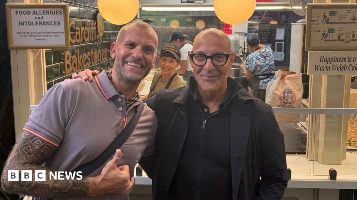 Owner of the Welsh cake store stood next to actor Stanley Tucci. He has a full sleeve tattoo and a bag strap across his body. Tucci is wearing a black jacket over a black quarter zip jumper. There is a staff member smiling in the background with a stack of Welsh cakes on the right. They sit under as sign reading: Happiness is a Warm Welsh Cake