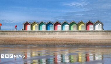 Blyth beach at low tide, looking back to the beach from the water. Colourful huts line the beach and a man with a red top runs along the promenade. The huts are reflected in the water and the sky is very blue.