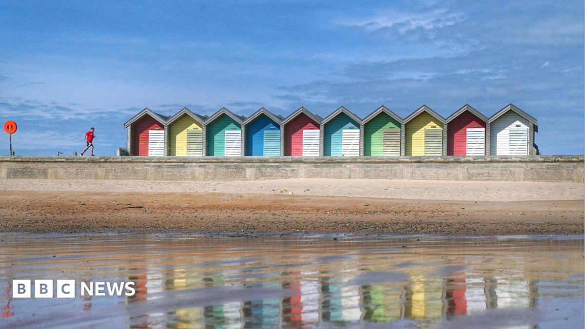 Blyth beach at low tide, looking back to the beach from the water. Colourful huts line the beach and a man with a red top runs along the promenade. The huts are reflected in the water and the sky is very blue.